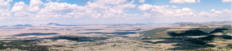 [Multiple photos stitched together creating a vista of the plains 2000 feet below the volcano rim. There are mountains in the distance and many puffy white clouds in the sky which provide shadow on certain sections of the plains.]